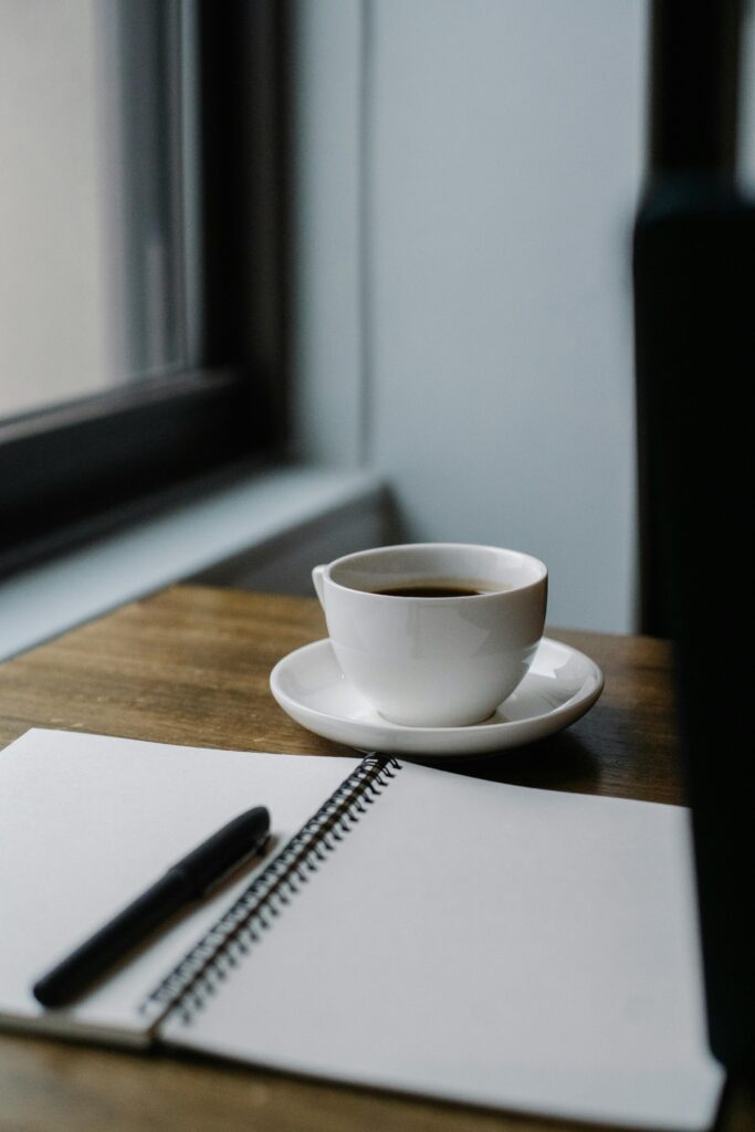 A cup of coffee and an open notebook by a window in a cozy café setting.